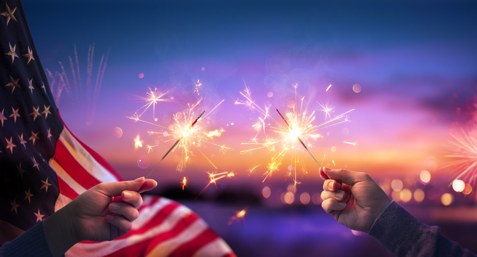 Hands holding sparklers against a background of fireworks, for a blog post from LCA on fireworks safety.