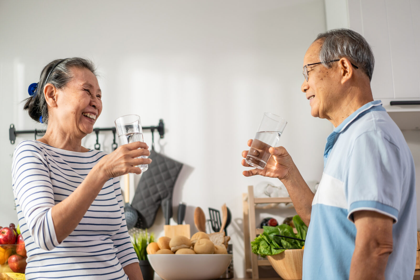 An Asian senior couple drink a glass of tap water in the kitchen. The photo illustrates an LCA post on Drinking Water Week.