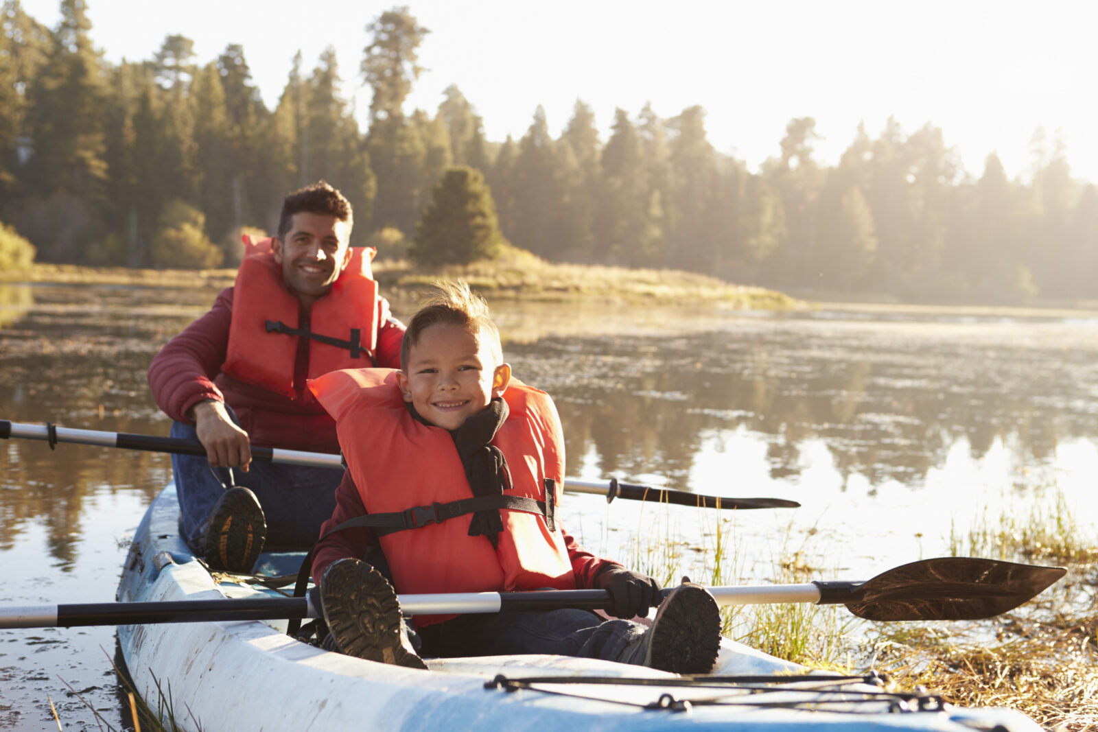 A father and son, wearing bright orange life jackets, enjoy a safe afternoon on a lake in a kayak.